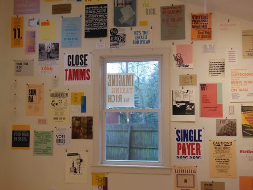 A series of multicolored printed posters displayed around a window. Some posters have a design, others have phrases like “Single Payer, Now! Now!” and “We are not born equal. We become equal in struggle.” Some reference political issues, other reference musicians, all feature graphic design