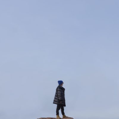Girl in black jacket standing on rock looking over her shoulder at expansive blue sky