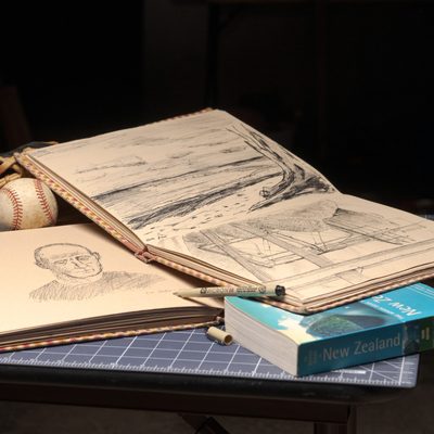 Two sketchbooks, an old New Zealand travel book, and a baseball glove are arranged on top of art cutting boards