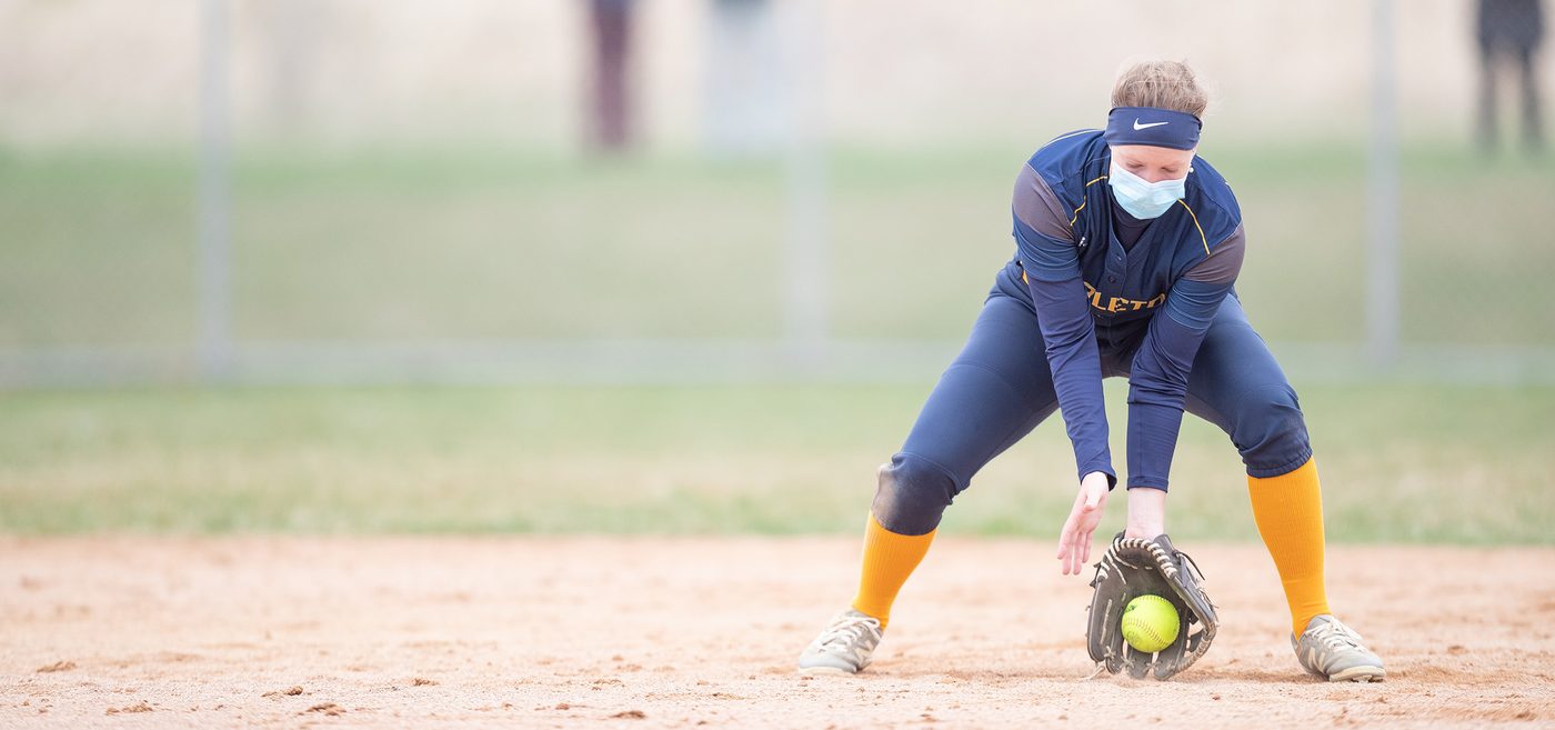 A softball player wearing a medical mask catches a ground ball