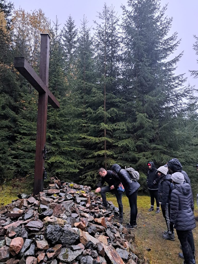 In an evergreen forest, the guide lights a candle next to a wooden cross with a pile of rocks in front of it.