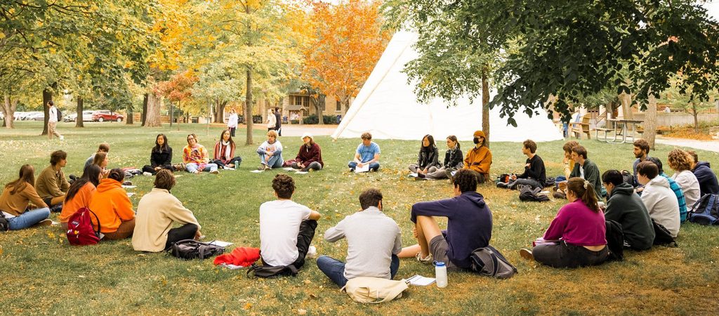A group of students sitting in a circle with a tipi in the background