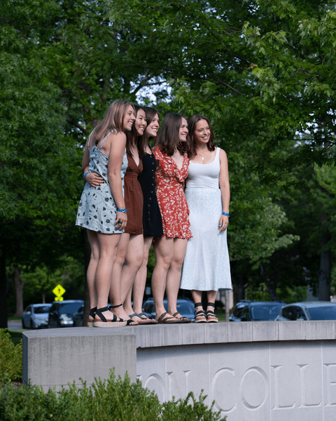 Five members of the Class of 2022 pose for a photo while standing on the Carleton College sign