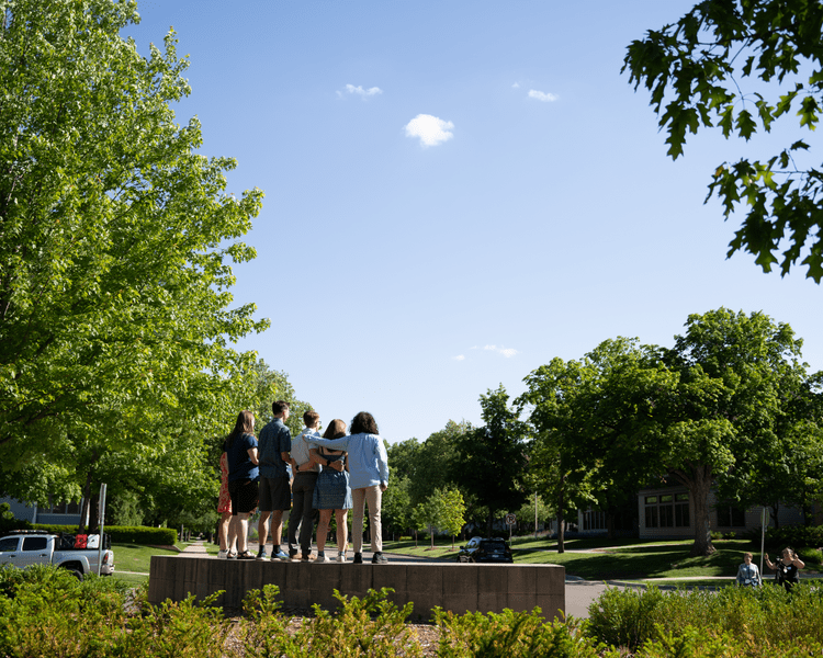 Five members of the Class of 2022, viewed from the back, stand on the Carleton College sign