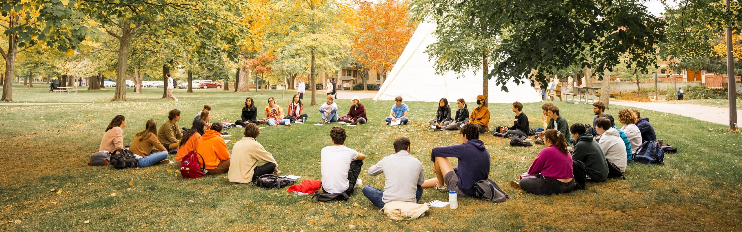 A class meets outside sitting in a circle, with a native American tipi in the background