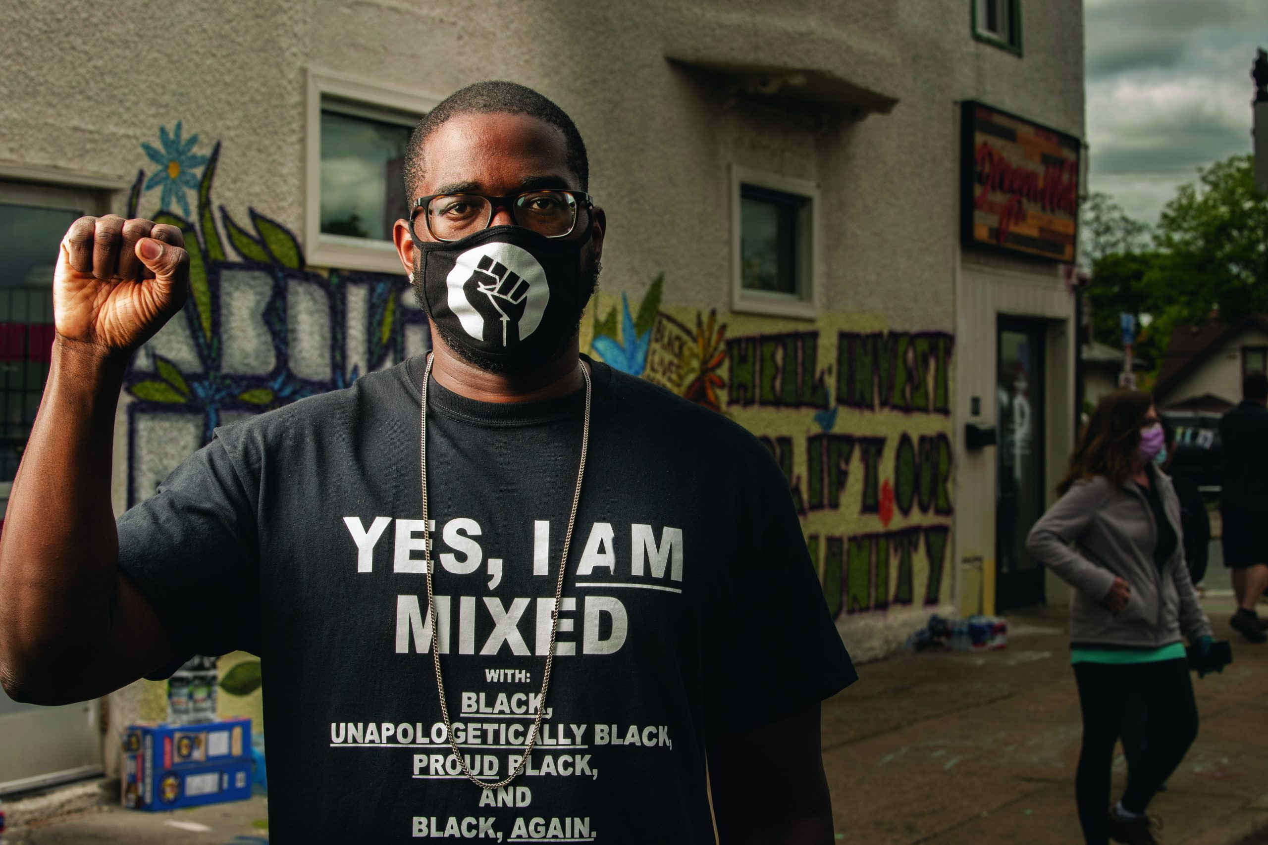 A Black man raises his fist. He wears a facemask with an illustration of a raised fist, and a T-shirt that reads: Yes, I am mixed with Black, unapologetically Black, proud Black, and Black, again
