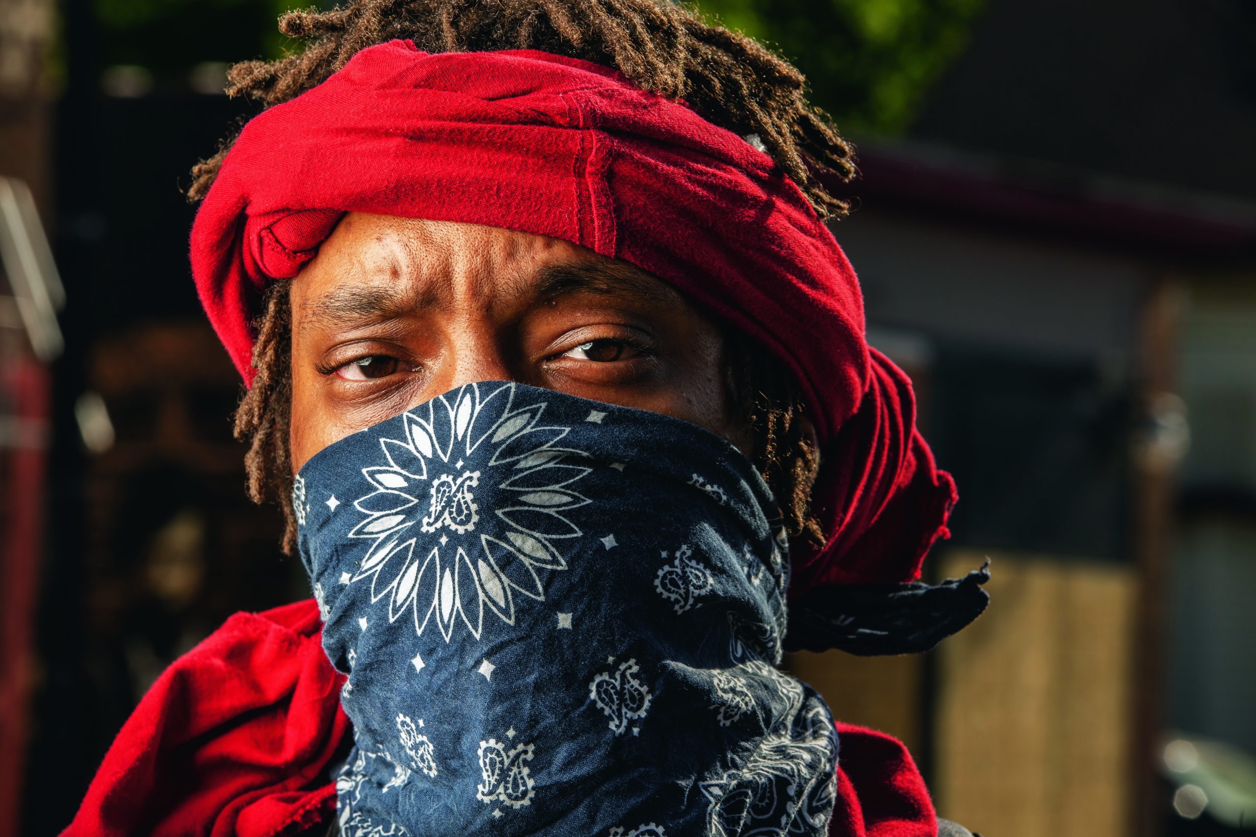 A dark-skinned man with a red cloth wrapped around his dreadlocked hair and a blue bandana covering his face