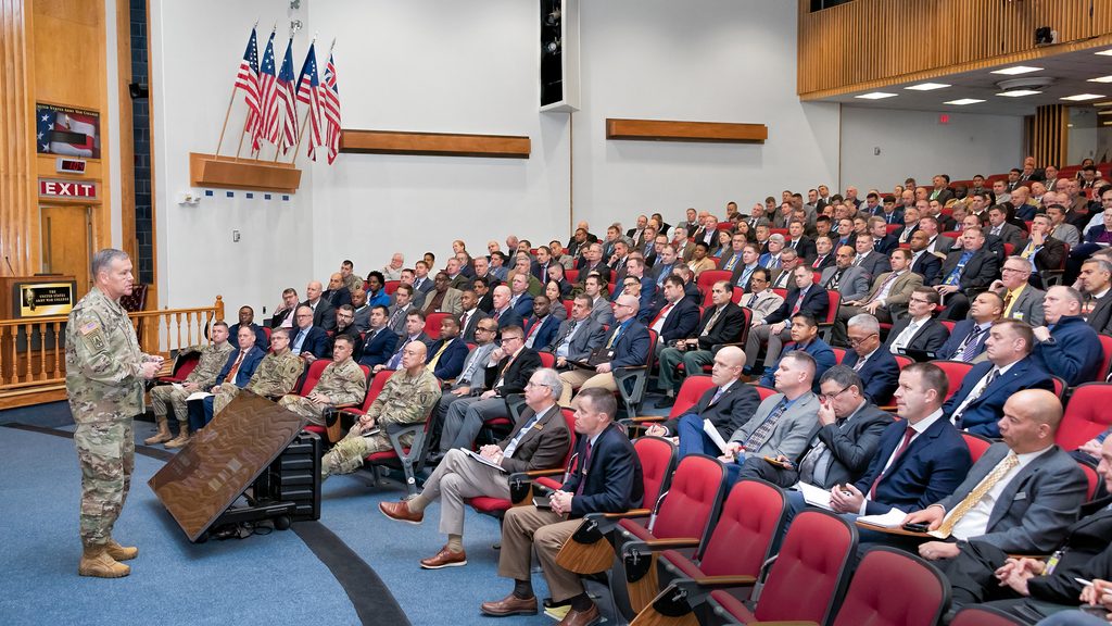 A man in military fatigues speaks to an audience of men wearing suits