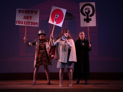 Three women stand in a triangular formation holding protest signs. The woman in the front of the trio also has a megaphone. One of the signs held by a woman in back reads “Women’s Art Coalition” in red and black block capital letters.