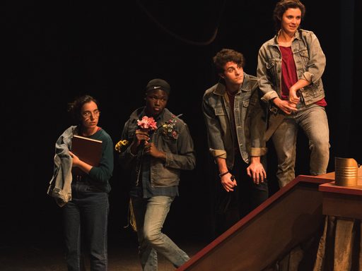 Four people, all wearing jeans and jeans jackets, are lined up on a stairway, looking stage left with varying expressions on their faces.