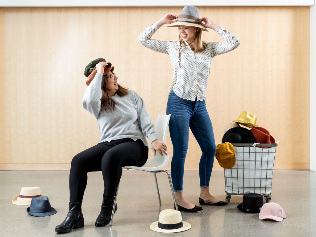 Two students pose with a basket of different hats, spilling onto the floor