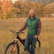A man stands in a meadow beside a bicycle with a mountain in the background