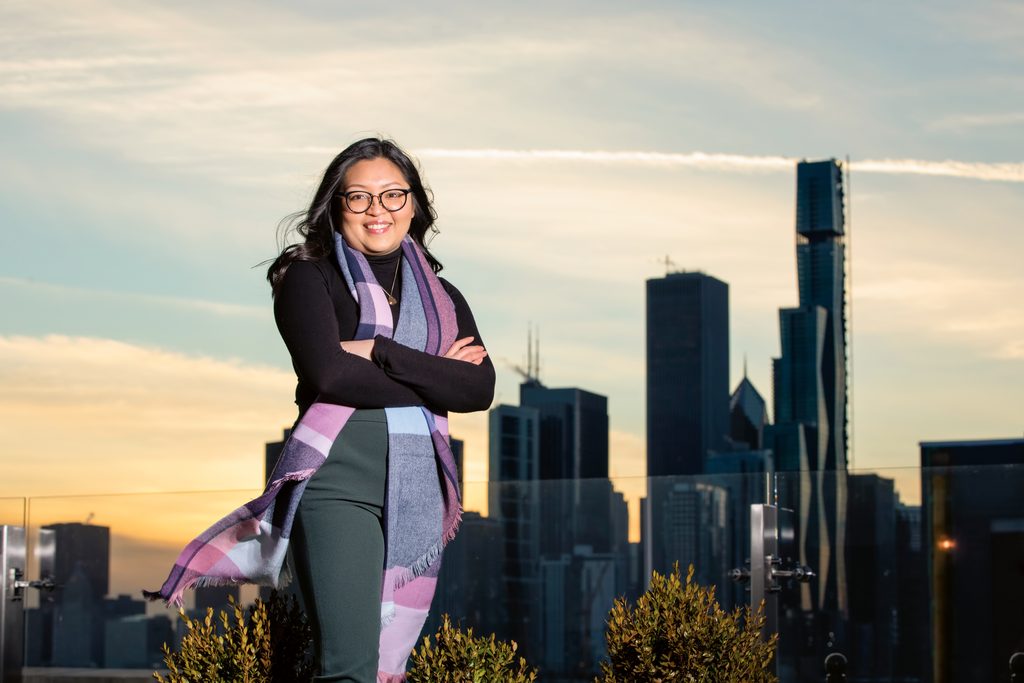 Lilly Athamanah ’15 smiles and poses with her arms crossed in front of tall buildings